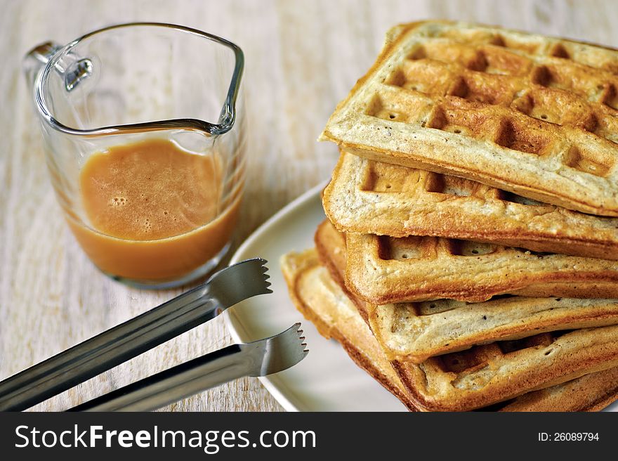 Stack of homemade waffles on the plate with a cup of brown syrup and cooking tong
