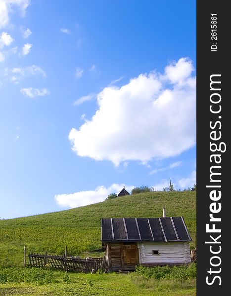 Background on a rural theme. In a photo a summer landscape with a green grass, the blue sky and an old log hut