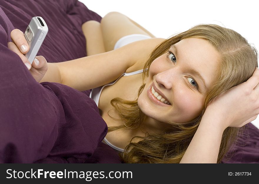 Young, beautiful woman lying in bed on violet coverlet. Smiling and holding mobile phone. Looking at camera. White background. Young, beautiful woman lying in bed on violet coverlet. Smiling and holding mobile phone. Looking at camera. White background