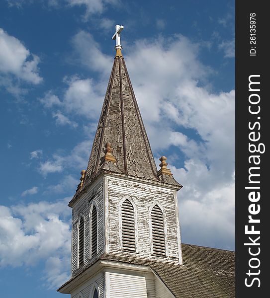 Close-up of an old, worn, and weathered wooden Christian church steeple with a cross on top, against a blue sky with clouds. Close-up of an old, worn, and weathered wooden Christian church steeple with a cross on top, against a blue sky with clouds.