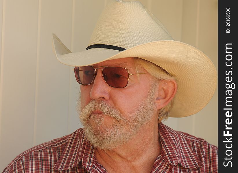 An older bearded man with a white cowboy hat, checked shirt and dark glasses is looking off to the side, with a serious concerned look. An older bearded man with a white cowboy hat, checked shirt and dark glasses is looking off to the side, with a serious concerned look
