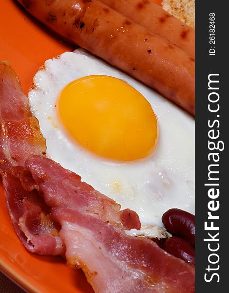 English Breakfast with Sausage, Bacon, Fried Eggs Sunny Side up and  Beans, close up on orange plate. Focus on Egg Yolk. English Breakfast with Sausage, Bacon, Fried Eggs Sunny Side up and  Beans, close up on orange plate. Focus on Egg Yolk