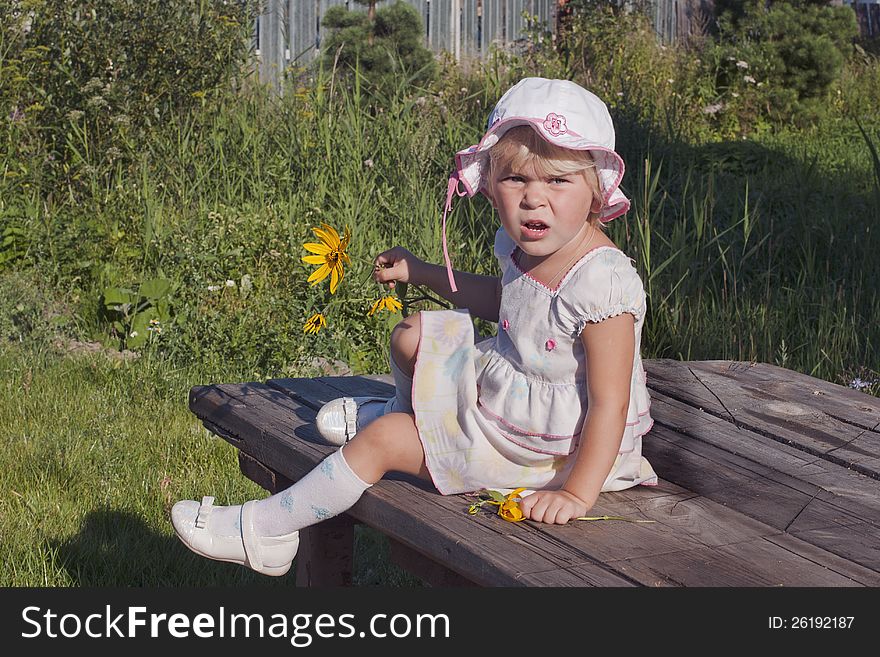 Happy child the girl in a white dress and a white cap with flower sitting on green grass outdoors in spring garden. Happy child the girl in a white dress and a white cap with flower sitting on green grass outdoors in spring garden