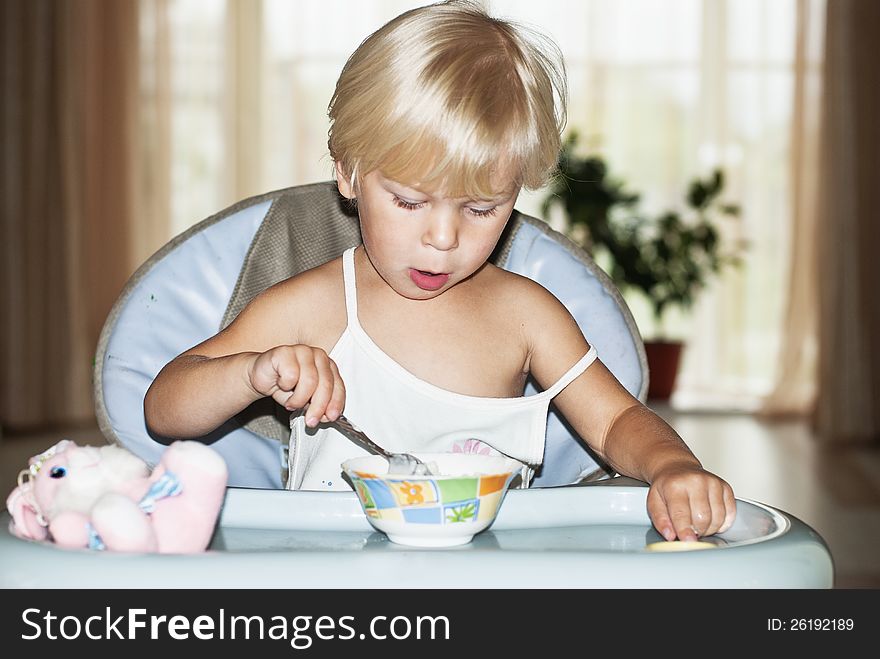 The nice girl (2 years), the blonde, sits at a little table and eats porridge. The nice girl (2 years), the blonde, sits at a little table and eats porridge