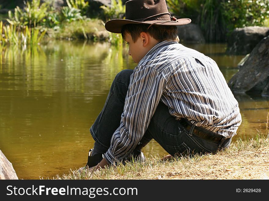 A child wearing jeans, shirt and hat sits quietly by the edge of a pond. A child wearing jeans, shirt and hat sits quietly by the edge of a pond