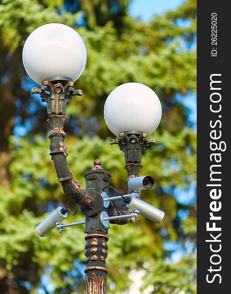 Security surveillance camera on a column with a lamp, on green tree background, selective focus