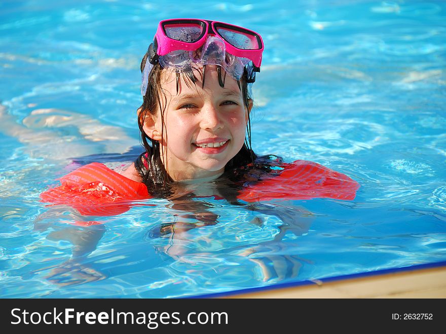 A young girl smiles into the camera as she takes a dip in the pool on a sunny day. She is wearing floats and goggles. A young girl smiles into the camera as she takes a dip in the pool on a sunny day. She is wearing floats and goggles.