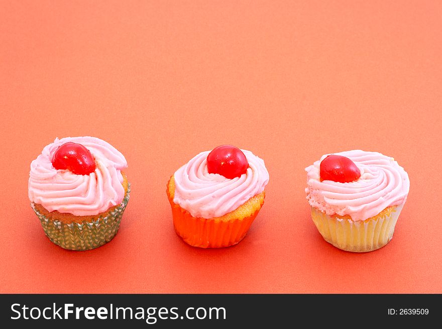 Three small cupcakes with cherries on orange background. Focus is on the middle cherry. Three small cupcakes with cherries on orange background. Focus is on the middle cherry