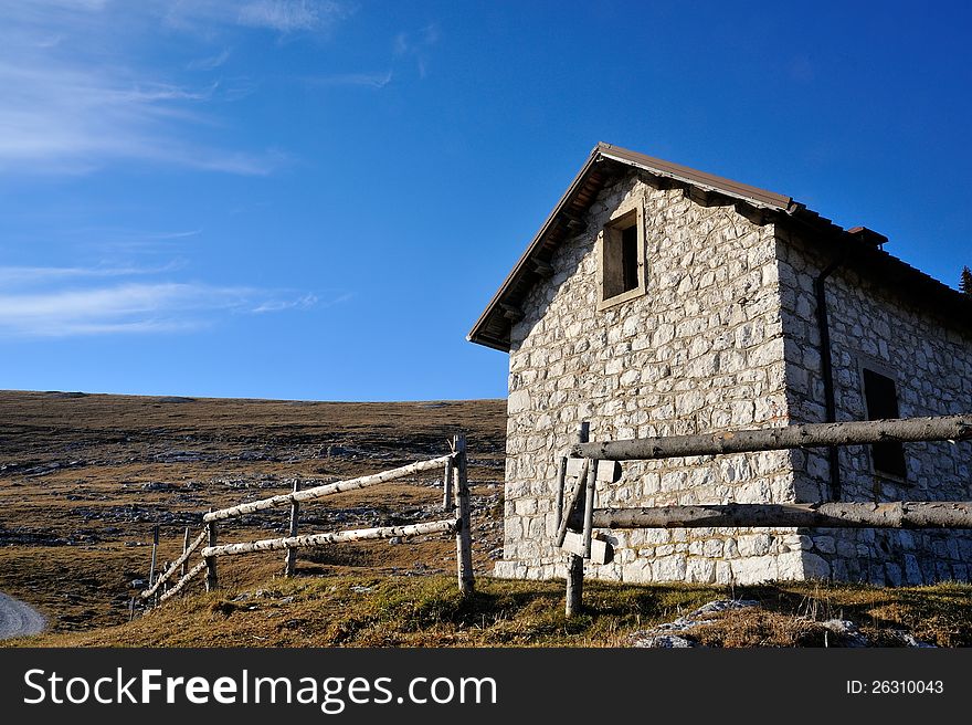 Old cabin in the mountain with stone walls, wooden fence in the green meadow and blue sky with clouds