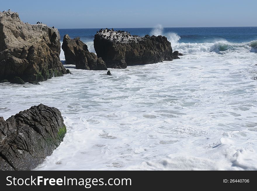Crashing waves on dark rocks with comorant birds on one of them with a blue sky background. Crashing waves on dark rocks with comorant birds on one of them with a blue sky background