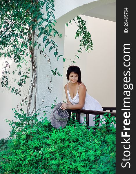 Girl in a white dress with black hat standing on the balcony and looking at a green tree. Girl in a white dress with black hat standing on the balcony and looking at a green tree