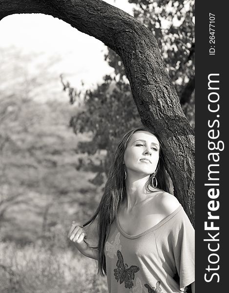 Black and white photograph of a pretty young brunette woman with long hair standing near a tree and enjoying the sun on a bright day. Black and white photograph of a pretty young brunette woman with long hair standing near a tree and enjoying the sun on a bright day