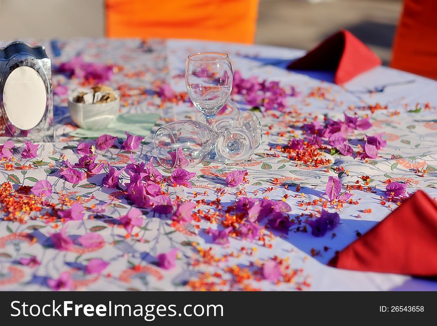 On the table and scattered flower petals are red wine glasses and napkins. On the table and scattered flower petals are red wine glasses and napkins