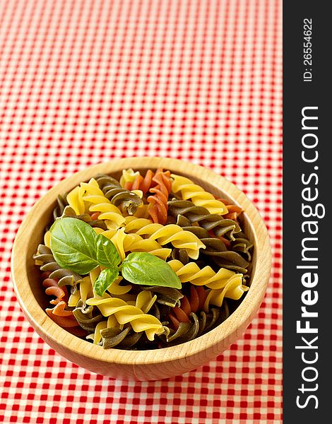 Multicolored pasta uncooked with a leaf of basil in a wooden bowl on red background