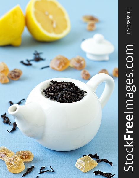 Small tea pot with leaves of black tea, brown crystal sugar and lemons on light blue background