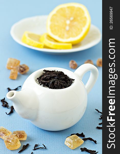Small tea pot with leaves of black tea, brown crystal sugar and lemons on light blue background