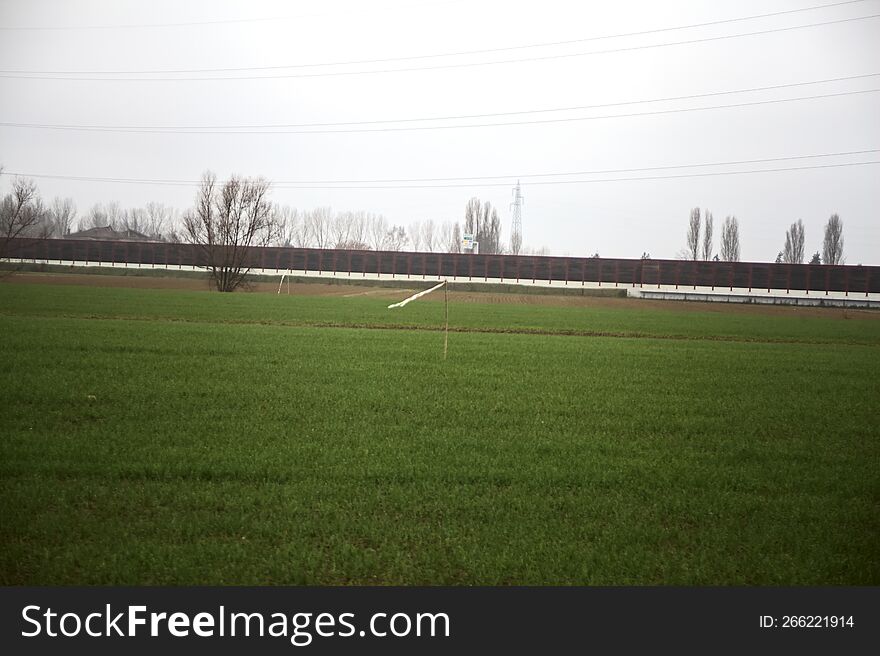 Cultivated field with trees in the distance framed by wooden pylons and over head cables on a cloudy day