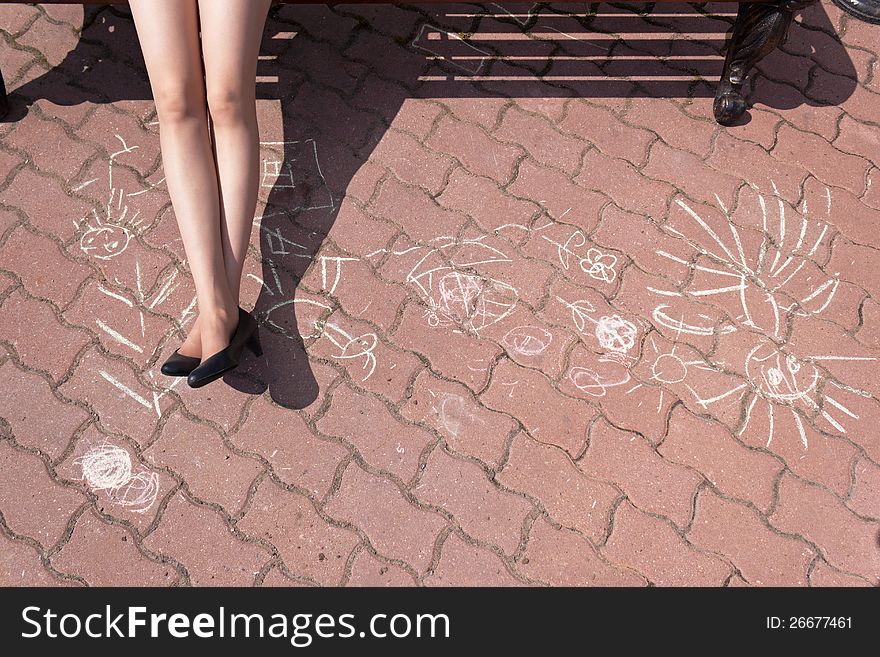 The girl sits in park on a bench, having extended beautiful feet. On sidewalk before a bench children have drawn a white chalk different drawings. The girl sits in park on a bench, having extended beautiful feet. On sidewalk before a bench children have drawn a white chalk different drawings.