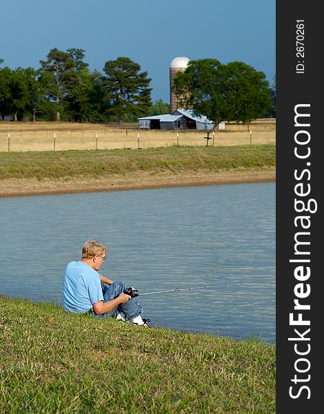 An older man sitting by the pond fishing with a pasture and old barn in the background. An older man sitting by the pond fishing with a pasture and old barn in the background.