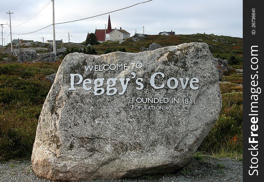 Welcome sign carved into huge boulder marking the site of Peggy's Cove and Lighthouse. Village buildings in in background. Welcome sign carved into huge boulder marking the site of Peggy's Cove and Lighthouse. Village buildings in in background.