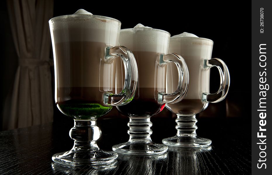 A group of three glasses of hot layered alcoholic cocktails, decorated with milk foam on black bar counter