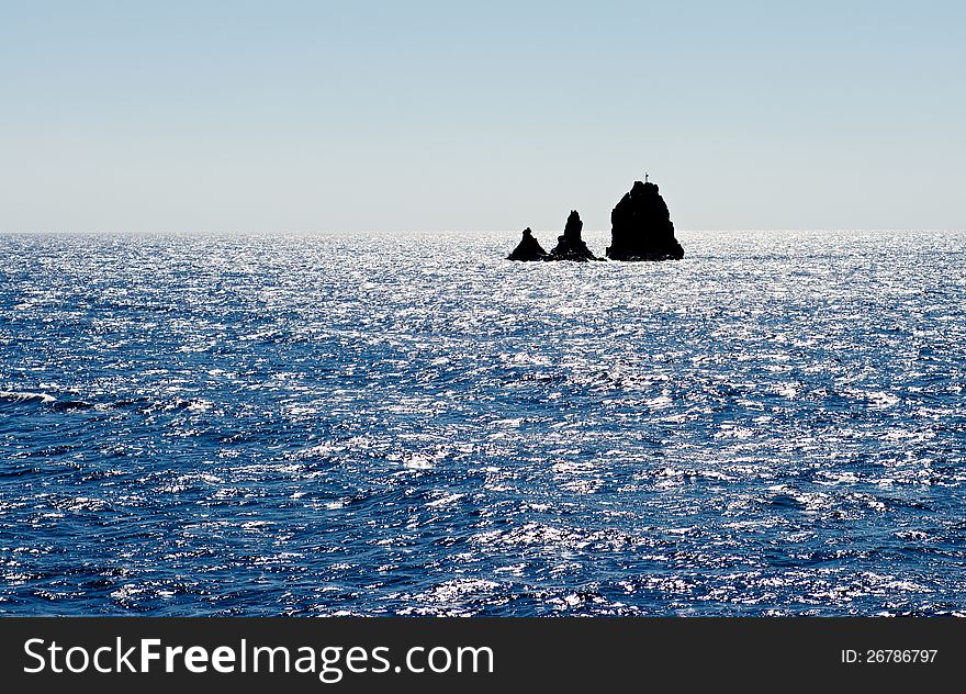 Black rocks are seen in the middle of the vast blue sea. Sea covered with patches of sunlight. Black rocks are seen in the middle of the vast blue sea. Sea covered with patches of sunlight.