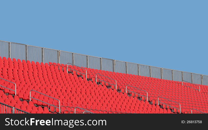 Section of empty red stadium seats in the top rows with a railing. against a blue sky with room for copy and text. Section of empty red stadium seats in the top rows with a railing. against a blue sky with room for copy and text.