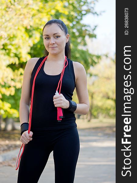 Athletic woman standing in a park in her sportswear with a skipping rope dangling around her neck. Athletic woman standing in a park in her sportswear with a skipping rope dangling around her neck