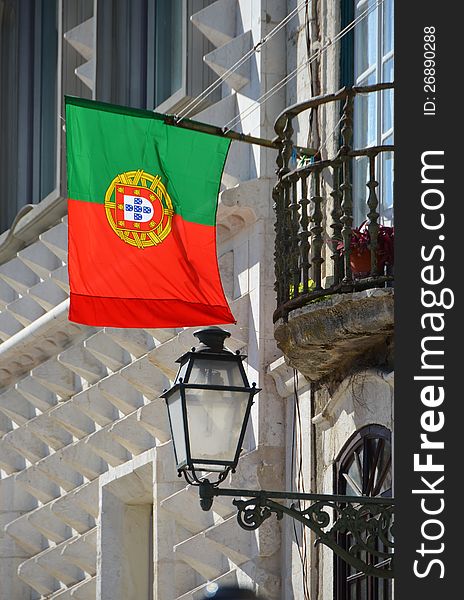 The flag of Portugal hangs from a house facade together with a street lamp and next to a balcony. The flag of Portugal hangs from a house facade together with a street lamp and next to a balcony.