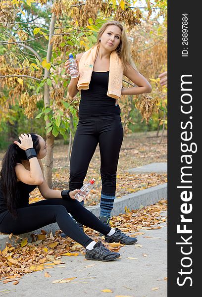 Two female athletes resting and drinking water sitting on the side of a rural road in their sportswear rehydrating after a heavy training session