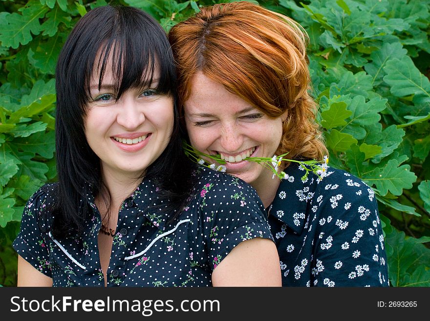 Two laughing girls on background of oak leaves. One girl keeps flower with teeth. Two laughing girls on background of oak leaves. One girl keeps flower with teeth