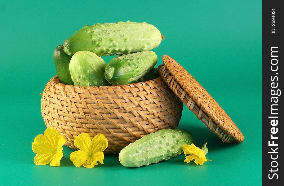 Some cucumbers lay in a basket. Beside the cucumber and flowers a cucumber lays. A green background. Some cucumbers lay in a basket. Beside the cucumber and flowers a cucumber lays. A green background.