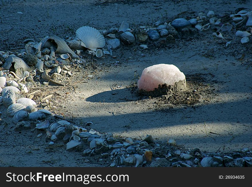 A large piece of rose quartz is in the center of a shell and stone circle outdoors, the quartz is casting a shadow. A large piece of rose quartz is in the center of a shell and stone circle outdoors, the quartz is casting a shadow.