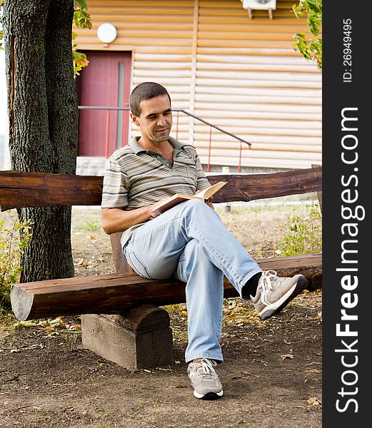 Man sitting relaxing reading a book on a rustic wooden bench in front of a timber log cabin. Man sitting relaxing reading a book on a rustic wooden bench in front of a timber log cabin