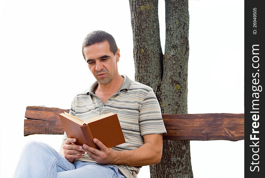 Middle-aged man reading a book sitting on a rustic wooden bench againsta tree trunk isolated on white