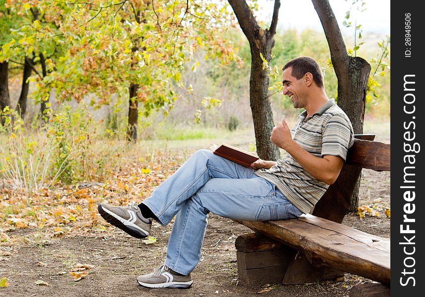 Middle-aged man sitting on a rustic wooden bench in the countryside laughing and reading as he thoroughly enjoys his book. Middle-aged man sitting on a rustic wooden bench in the countryside laughing and reading as he thoroughly enjoys his book.