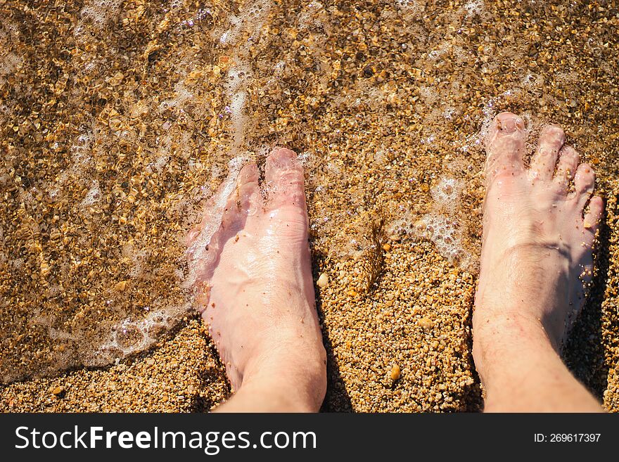 Men s feet are standing on a sandy beach. Small waves are foaming on the seashore. Crystal water.