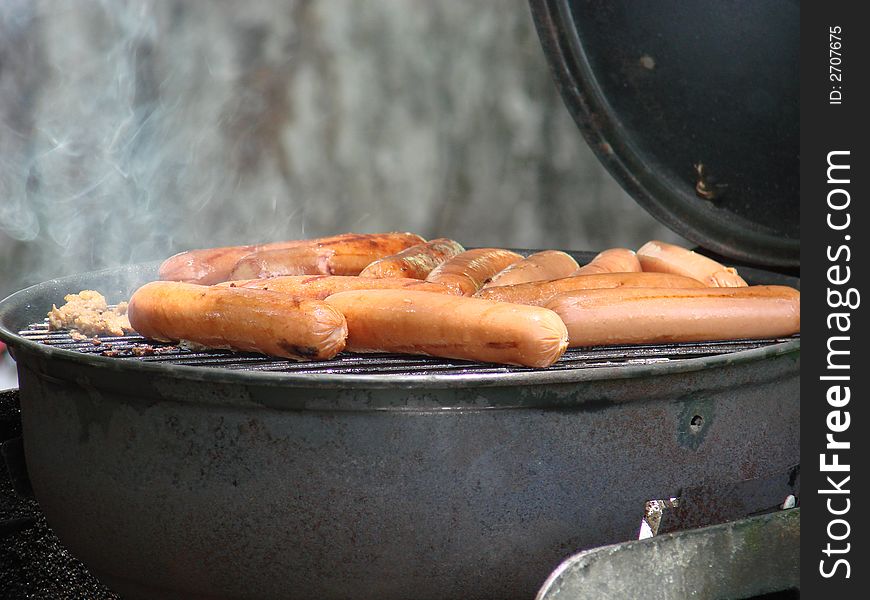 Lots of hot dogs cooking on a old grill with smoke rising.