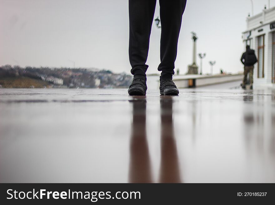 A man stands on wet asphalt in the rain. A park on the seashore. A muddy reflection on the ground.
