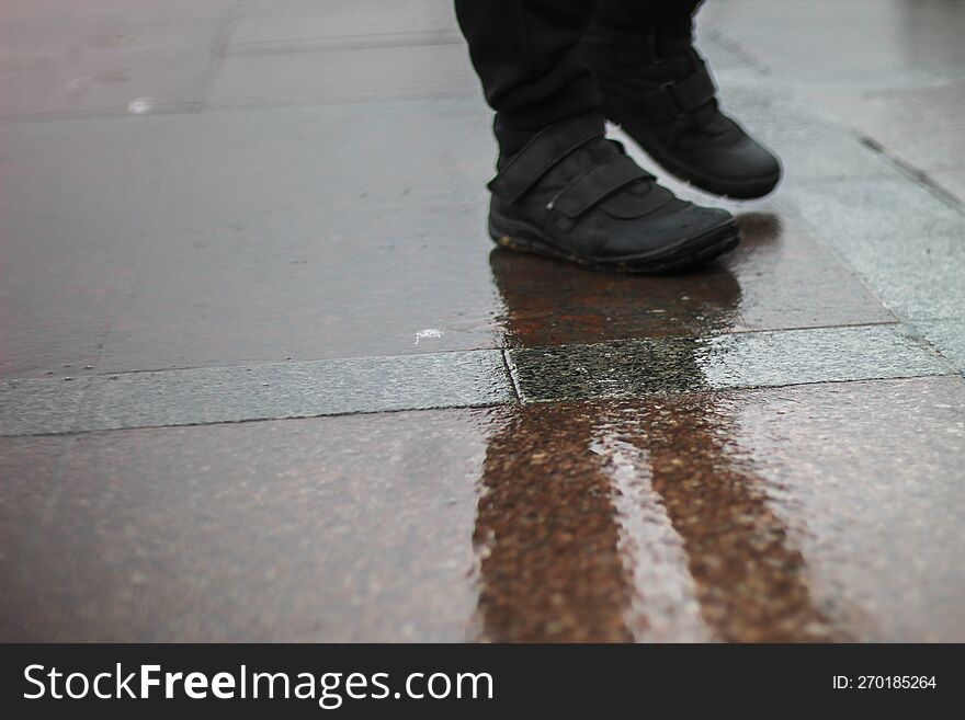 A Man Stands On Wet Asphalt In The Rain. A Park On The Seashore. A Muddy Reflection On The Ground.