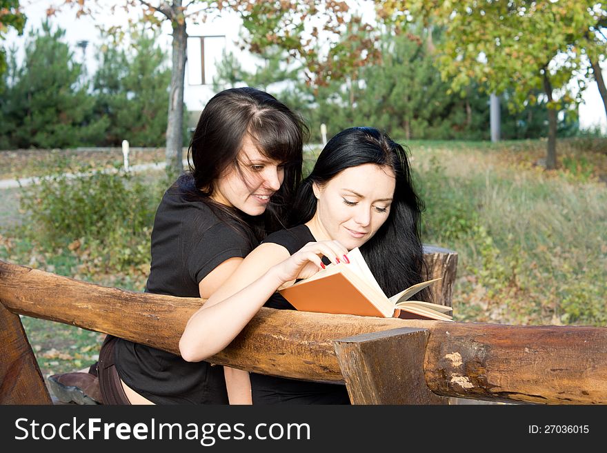 Two attractive young girls sitting on a rustic wooden bench reading a book together. Two attractive young girls sitting on a rustic wooden bench reading a book together