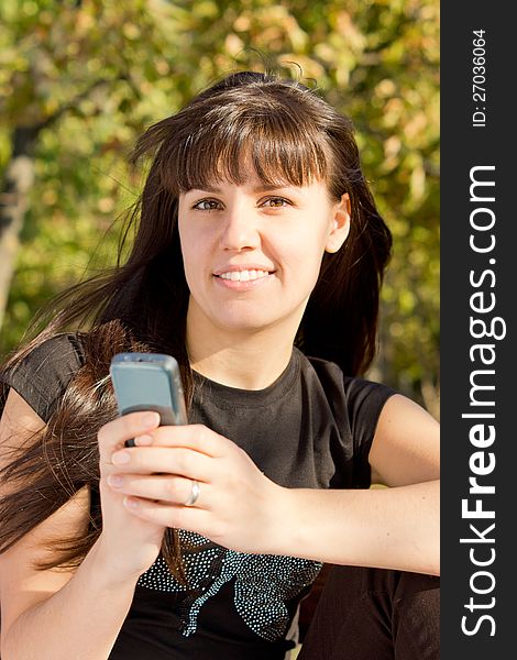 Woman holding her mobile phone sitting in a park and smiling. Woman holding her mobile phone sitting in a park and smiling.