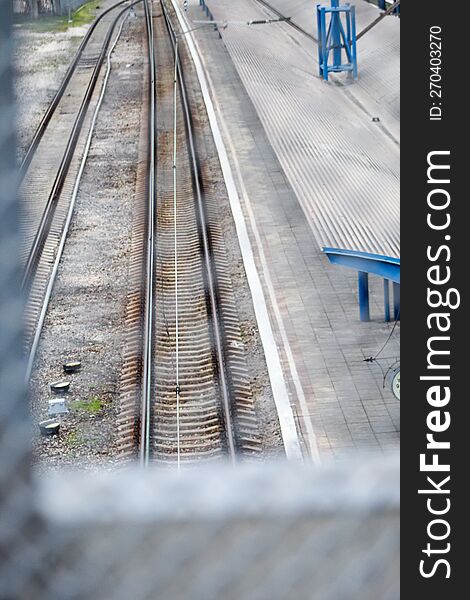 View Through The Metal Fence To The Empty Platform Of The Railway Station. Blue Columns At The Railway Station.