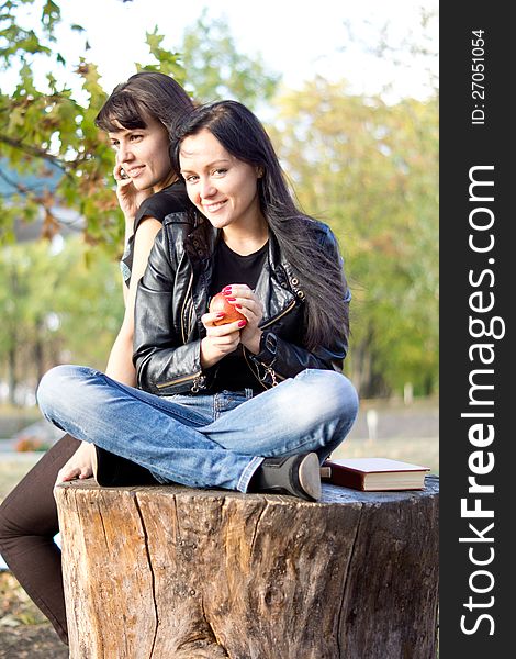 Young women seated on a sawn off tree trunk outdoors eating an apple while her friend chats on her mobile phone. Young women seated on a sawn off tree trunk outdoors eating an apple while her friend chats on her mobile phone