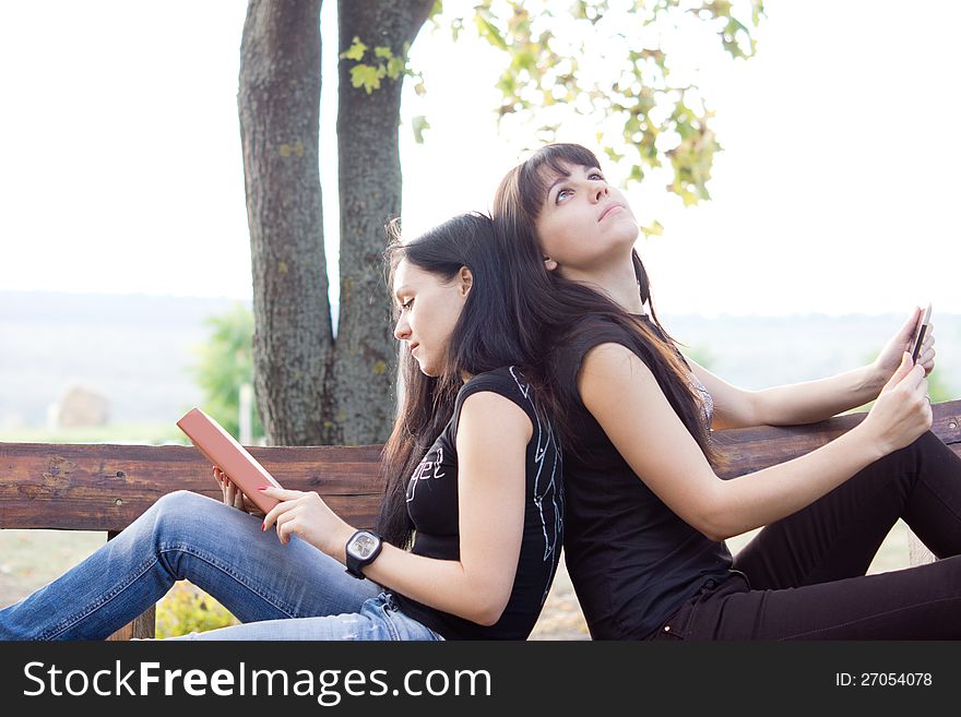 Two young female friends relaxing on a bench, one reading a book with the other stares thoughtfully into the sky while holding a tablet in her hands. Two young female friends relaxing on a bench, one reading a book with the other stares thoughtfully into the sky while holding a tablet in her hands
