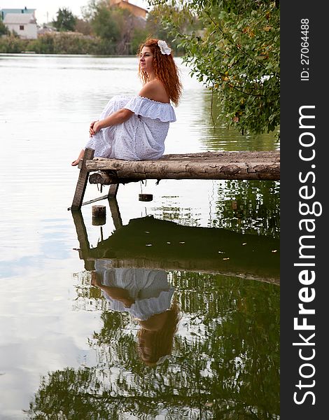 Beautiful young red-haired woman in white dress with flower in her hair or bride or undine sitting on a wooden bridge on the lake. Beautiful young red-haired woman in white dress with flower in her hair or bride or undine sitting on a wooden bridge on the lake