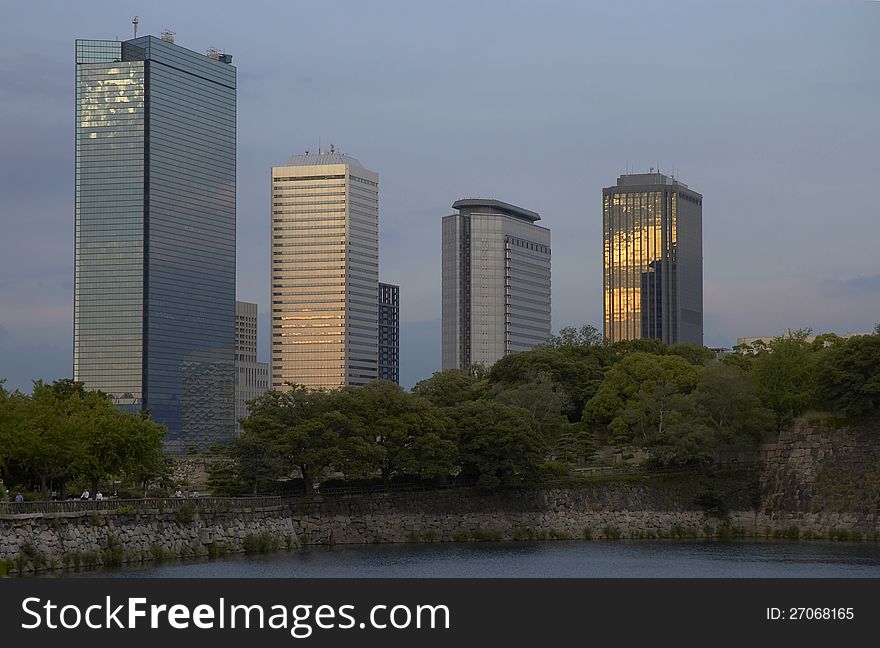 Skyscrapers with golden reflections at sunset behind a small forest with water