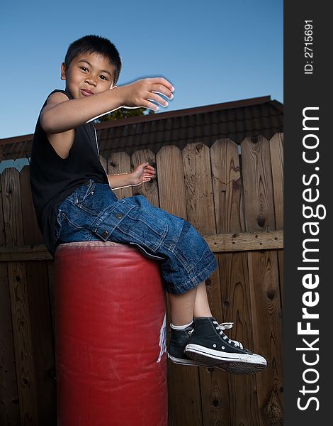 Young asian boy sitting on top of a punching bag outside beside a tall wooden fence smiling wearing jeans and black tshirt. Young asian boy sitting on top of a punching bag outside beside a tall wooden fence smiling wearing jeans and black tshirt