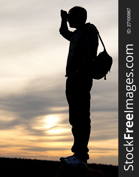 Silhouette of a man with a satchel on his back standing with his hand raised to his forehead looking into distance against a delicate sunset. Silhouette of a man with a satchel on his back standing with his hand raised to his forehead looking into distance against a delicate sunset