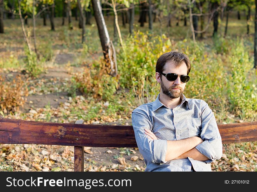 Young bearded man sitting with his arms folded on a rustic wooden bench deep in thought. Young bearded man sitting with his arms folded on a rustic wooden bench deep in thought